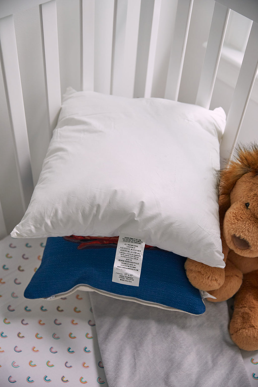 A white-framed crib holds two square Sewing Down South 17 x 17 Indoor Nursery Inserts filled with non-allergenic fiber, a brown lion plush toy, and a folded gray blanket on a patterned fitted sheet. Natural light streams in through a nearby window.