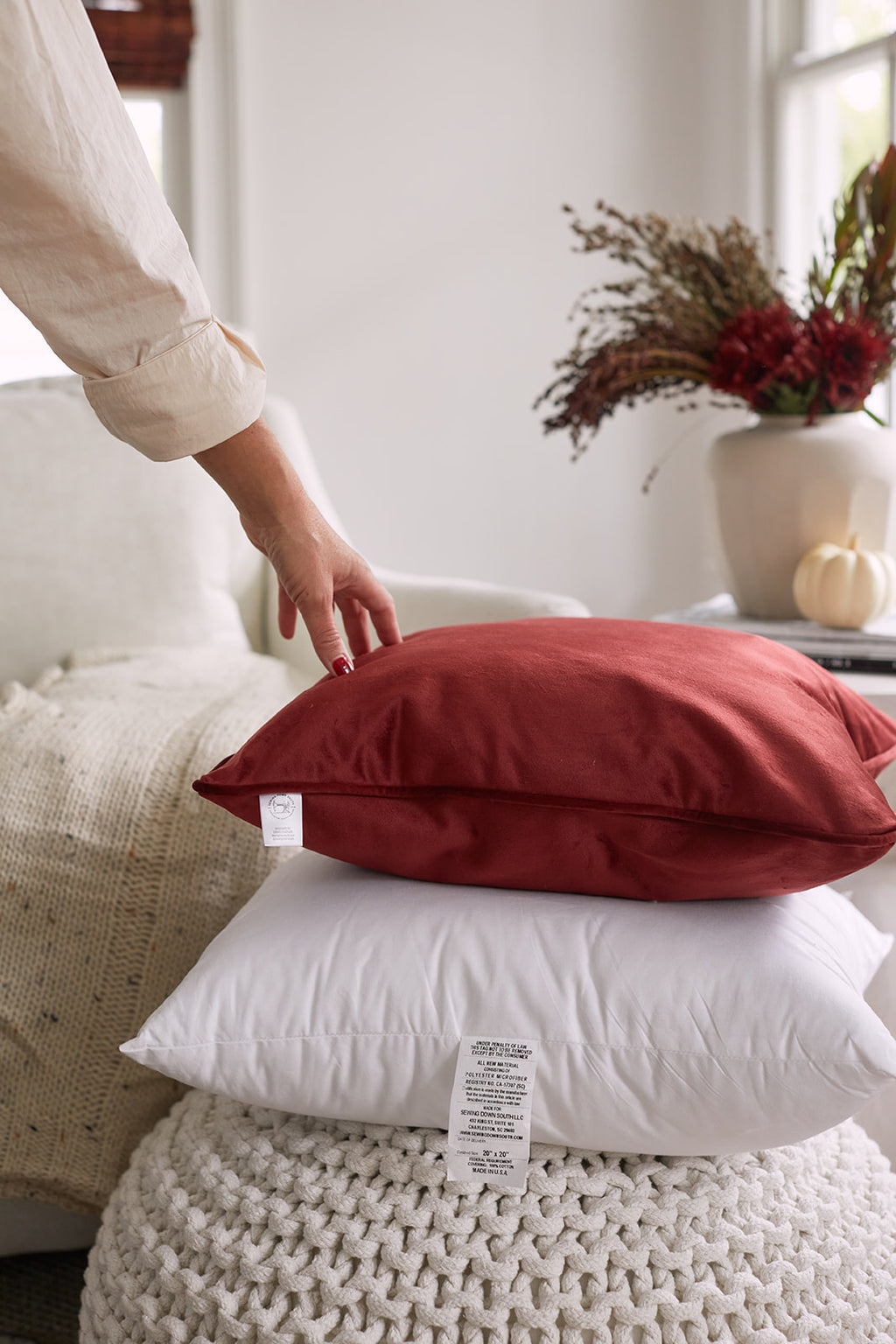 Two Sewing Down South 20 x 20 white pillows filled with non-allergenic fiber rest on a beige sofa next to a knitted beige blanket. Bamboo blinds softly filter the light through the window, creating a cozy and inviting atmosphere.