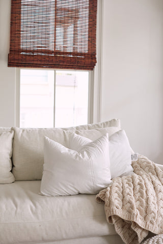 The Sewing Down South 16 x 20 Indoor Inserts add premium comfort with lumbar support, while a beige knitted blanket complements the white sofa. Soft light through partially open wooden blinds creates a cozy, minimalist vibe.