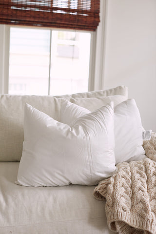 A cozy scene includes two white Sewing Down South 16 x 20 Indoor Inserts and a premium lumbar pillow on a beige sofa, paired with a knitted blanket. Sunlight passes through window blinds, casting a soft glow and enriching the tranquil ambiance.