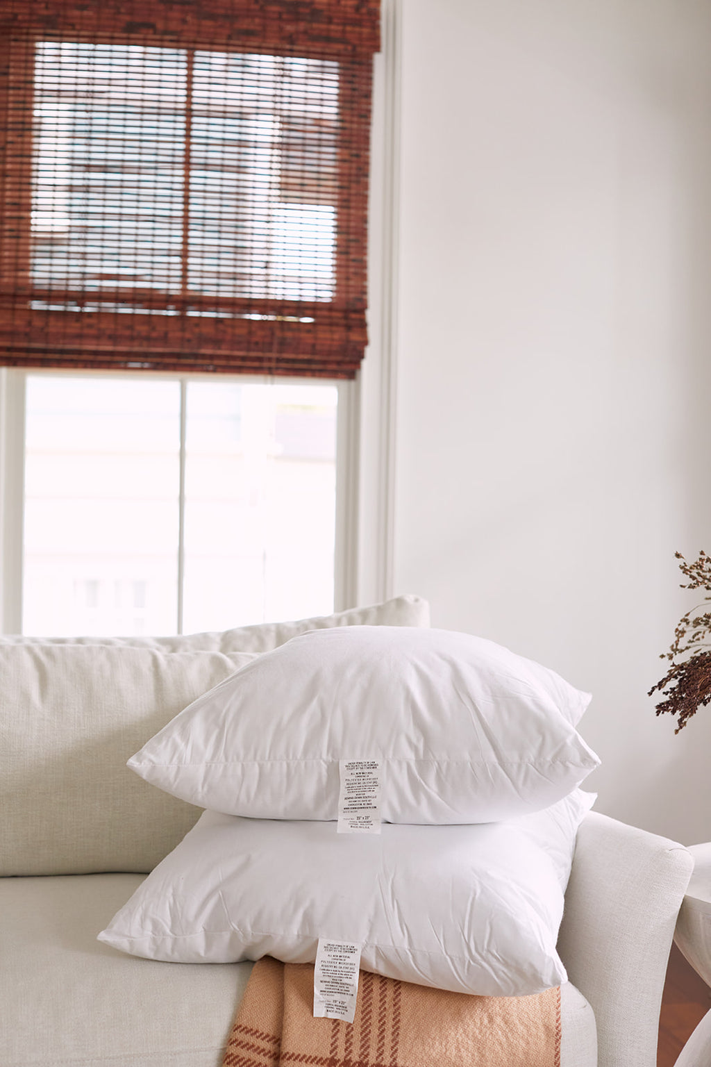 A cozy living room scene showcases a white sofa with two pillows: one large using a Sewing Down South 23 x 23 Indoor Insert and a smaller orange pillow with a leaf pattern. Nearby, a plaid blanket is draped, and bamboo shades on the window are partially pulled down.