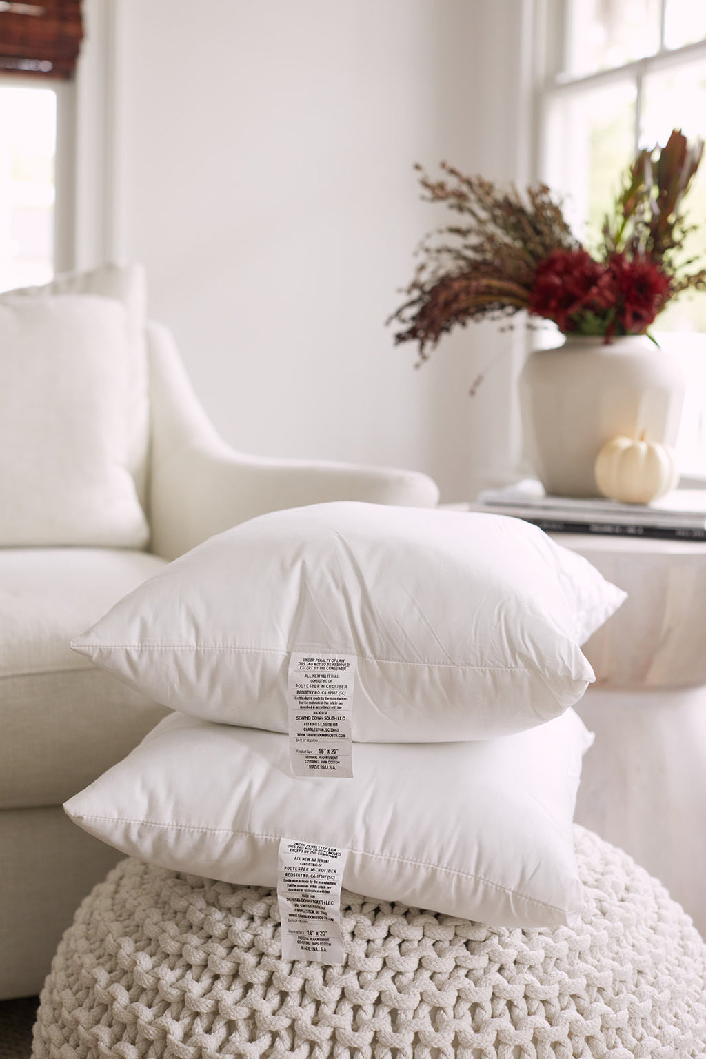 A cozy scene includes two white Sewing Down South 16 x 20 Indoor Inserts and a premium lumbar pillow on a beige sofa, paired with a knitted blanket. Sunlight passes through window blinds, casting a soft glow and enriching the tranquil ambiance.