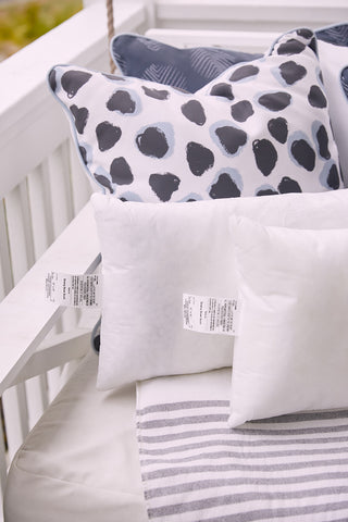 Two Sewing Down South 16 x 20 Outdoor Inserts, filled with mold-resistant polyester cluster fiber, rest on a cushion beside a gray-striped blanket. Decorative spotted pillows are arranged behind them on a white outdoor bench or swing.