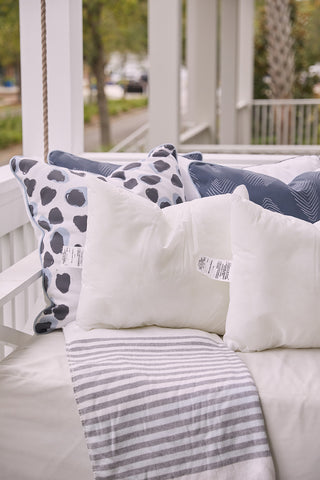 A cozy outdoor porch swing with white and blue patterned pillows—two featuring the Sewing Down South 16 x 20 Outdoor Insert with polyester cluster fiber fill—and a white blanket with gray stripes, set against greenery and a white railing.
