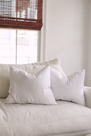 A cozy white sofa with premium lumbar pillows, 16x20 Sewing Down South Indoor Inserts, offering style and support. Two fluffy inserts add comfort as wooden blinds filter soft natural light through the window, creating an inviting atmosphere.