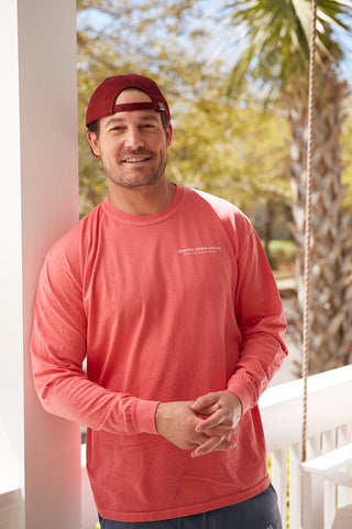 A man in a vintage-looking red cap and the Sewing Down South Long-Sleeve Logo Tee in Rainbow Row Colors leans against a white porch railing, smiling comfortably. The sunny backdrop with palm trees and lush greenery enhances the relaxed vibe, while his 100% ring spun cotton tee adds to the comfort of the scene.