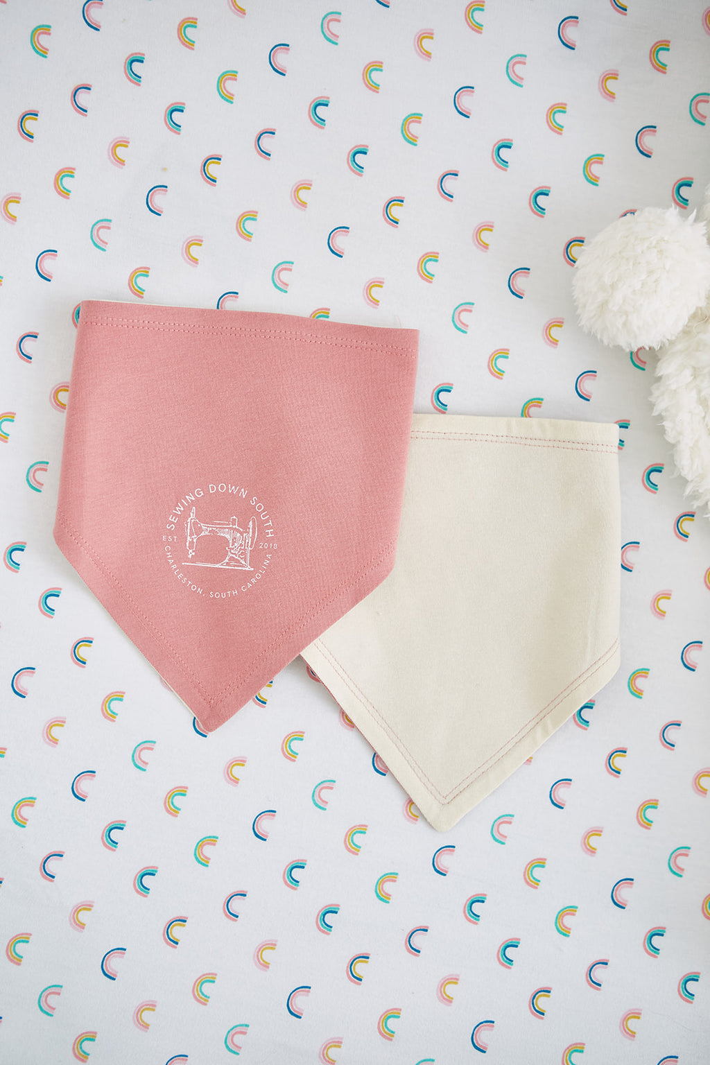 Two SDS Bandana Bibs, one light blue with a sewing machine logo and one dark blue, lie on rainbow-patterned fabric. Ideal for a baby gift, the scene is made even more charming by a white plush teddy bear peeking in from the right.