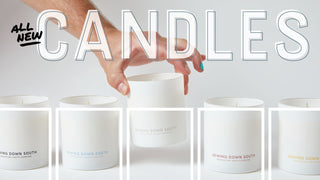 A hand lifts a white candle from a row of five candles in white jars labeled Sewing Down South. Large text at the top reads ALL NEW CANDLES on a white background.