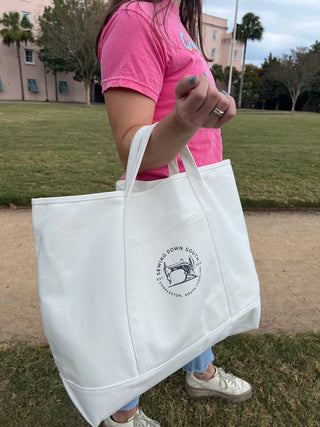 A woman wearing a pink shirt holds a large white tote bag with the Sewing Down South logo on it, standing outside on a grassy area with buildings and trees in the background.