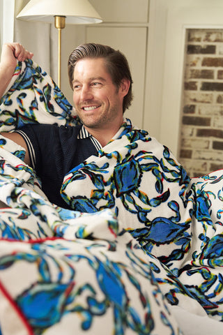 A smiling man sits wrapped in a white blanket with a colorful blue crab pattern, in a cozy room with a lamp and a brick wall in the background.