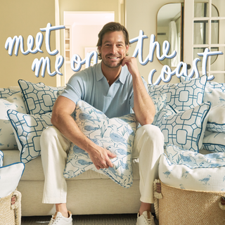 A smiling man sits on a sofa surrounded by blue and white patterned pillows, holding one on his lap. Behind him, text reads, meet me on the coast. The scene is bright and inviting.
