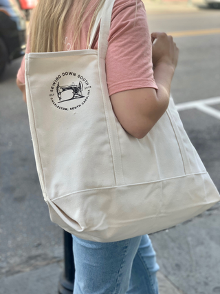 A person with long blonde hair wearing a pink shirt and light blue jeans carries a large white tote bag with a sewing machine logo and text that reads Sewing Down South Charleston, South Carolina.