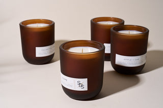 Four brown glass candles with white wax and minimal white labels are arranged on a light surface, each labeled with different scents such as Apple + Oak and Palmetto.