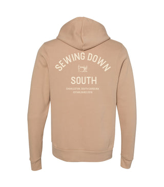 Tan hooded sweatshirt seen from the back, featuring white text that reads Sewing Down South, Charleston, South Carolina, Established 2018 with a small sewing machine graphic above the text.