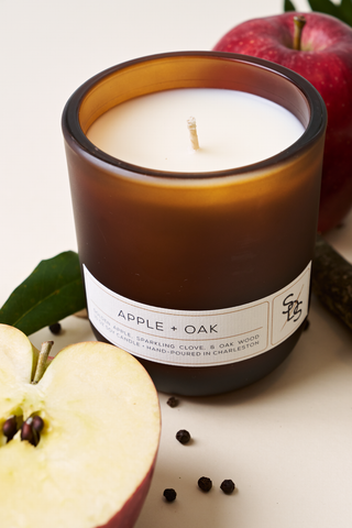 The Apple + Oak Signature Candle, a brown glass soy candle hand-poured in Charleston, is shown beside a whole red apple, sliced apple, green leaves, and black peppercorns on a light surface.