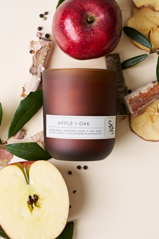The Apple + Oak Signature Candle, a brown glass soy candle hand-poured in Charleston, is displayed on a light surface with a whole red apple, sliced apple, oak bark, green leaves, and peppercorns.