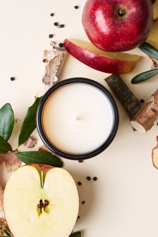The Apple + Oak Signature Candle, hand-poured in Charleston and housed in a black jar, is displayed with apples, leaves, wood pieces, and black peppercorns on a light surface to evoke its inviting apple oak scent.