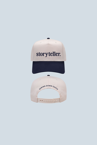 A Bravocon exclusive, the Storyteller 3D Embroidered Hat is a two-tone 5-panel cap with a white crown, navy blue brim, “storyteller.” embroidered on the front, and "SWING DOWN SOUTH" above the adjustable strap—all in black text.
