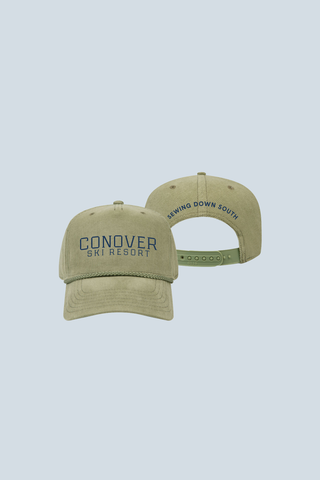 Two green Conover Ski Resort Embroidered Corduroy Hats sit on a light blue background. One cap has “CONOVER SKI RESORT” embroidered in blue, while the back reads “SEWING DOWN SOUTH” above the adjustable strap.