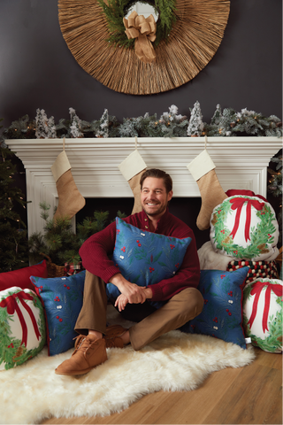 A man sits on a white rug in front of a decorated fireplace, holding the Holly Pillow. He is surrounded by festive decorative pillows, stockings, greenery, and a large wreath above the mantel.