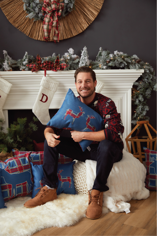 A man smiles while sitting on a white blanket by a festive fireplace, holding the Deer Pair Lumbar Pillow. The cozy scene includes Christmas stockings, greenery, and holiday accent pillows.