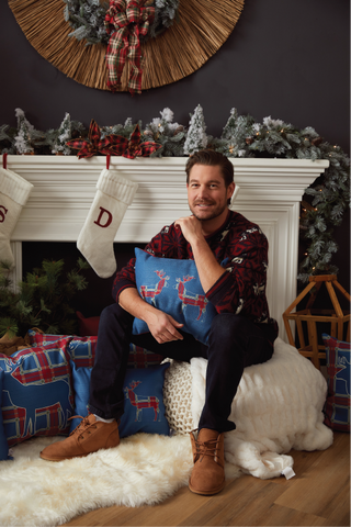 A smiling man sits on a fluffy white rug by a decorated fireplace, holding the Deer Pair Lumbar Pillow with red reindeer. Christmas stockings hang from the mantel, surrounded by festive holiday decorations.