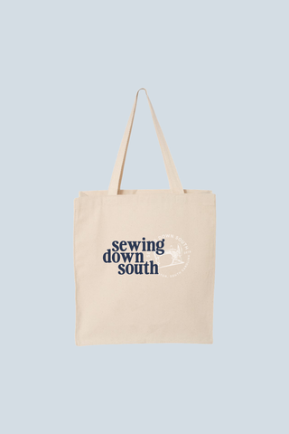 The SDS Brand Logo Tote is a beige cotton bag with two handles, featuring the bold navy phrase "Sewing Down South" and the SDS logo—a white circle with a sewing needle and thread—on a light blue background.
