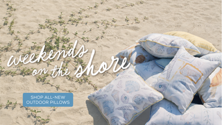 Outdoor pillows with coastal patterns arranged on a sandy beach, with the text 'weekends on the shore' and a blue badge reading 'SHOP ALL-NEW OUTDOOR PILLOWS'.