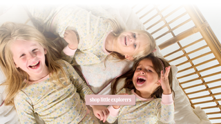 Three young girls lying on a bed wearing matching light yellow pajamas with a small floral pattern. A pink banner overlay reads 'shop little explorers'.