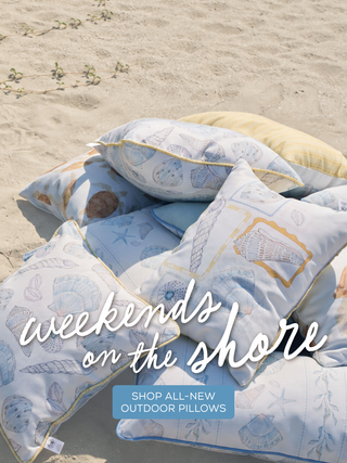 Outdoor pillows with coastal patterns arranged on a sandy beach, with the text 'weekends on the shore' and a blue badge reading 'SHOP ALL-NEW OUTDOOR PILLOWS'.