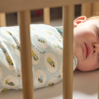 A sleeping baby wrapped in a light blue and white muslin swaddle blanket with a yellow and black bee pattern, lying in a wooden crib.
