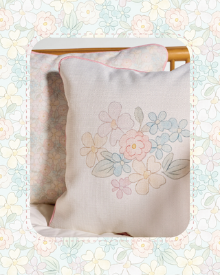 A square decorative throw pillow in cream linen fabric with a pastel embroidered floral design in pink, blue, and green, shown leaning against another pillow on a bed. The image is framed by a decorative floral border.