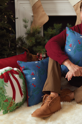 A person in brown pants and tan shoes sits on a white fluffy rug by a Christmas tree, beside a festive scarlet pillow, a blue pillow, and the Holly Pillow, which features a holiday wreath design. A white fireplace with stockings is behind them.