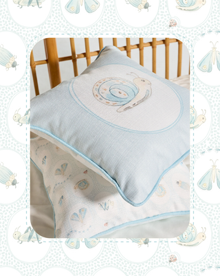 A light blue and white baby pillow with a snail embroidery, shown on a wooden crib with a matching patterned blanket.