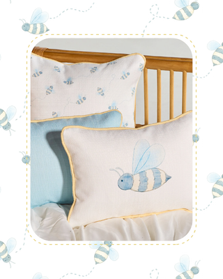 A styled nursery bedding set featuring a cream pillow with a blue and yellow bumblebee applique, a light blue pillow, and a cream pillow with a small bumblebee print, arranged on a wooden crib.