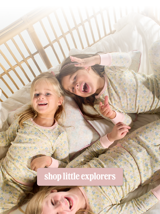 Three young girls lying on a bed wearing matching floral pajamas, with a pink banner overlay that reads 'shop little explorers'.