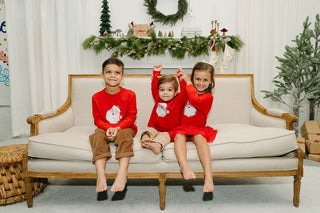 Three kids in matching Toddler & Childrens Santa Long Sleeve Tees (2T-XL) sit barefoot on a beige couch in a festive living room with Christmas greenery, stockings, and gifts. The middle child smiles with raised arms.