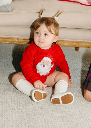 A baby with light brown hair in pigtails sits on a light carpet, wearing a Santa Baby Onesie with white socks and shoes, looking slightly to the side.