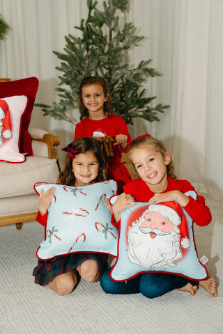 Three young girls in festive red tops sit by a Christmas tree, smiling and holding the Childrens Santa Pillow in Baby Blue from the Santa Holiday Pillow Collection. A beige chair with more holiday decor is beside them.
