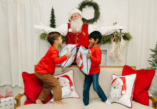 Two young boys in red shirts have a playful pillow fight on a festive couch with a man dressed as Santa Claus behind them, surrounded by holiday decor and the Childrens Santa Pillow in Baby Blue.