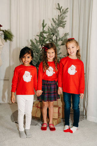 Three young girls stand in front of a sparkling Christmas tree, holding hands and smiling. One wears the Toddler & Childrens Santa Long Sleeve Tee (2T-XL), while presents sit wrapped beneath the festive scene.