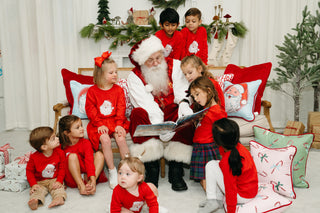 Santa Claus sits on a couch reading to children in festive red outfits, surrounded by holiday decor and wrapped presents. The joyful Christmas scene is enhanced by the Childrens Green Candy Cane Pillow.