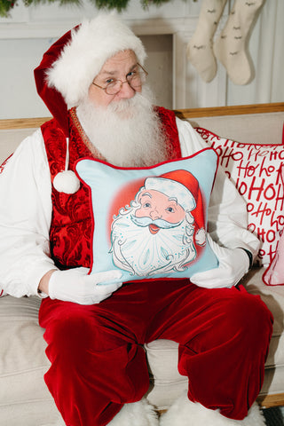 A man dressed as Santa Claus sits on a couch holding the Childrens Santa Pillow in Baby Blue, featuring a cartoon Santa face. Festive holiday decor and a cheerful Ho Ho Ho pillow are visible in the background.