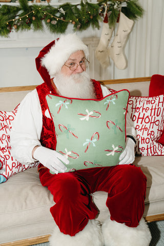 Santa Claus sits on a couch holding the Childrens Green Candy Cane Pillow, surrounded by festive Ho Ho Ho pillows and cheerful holiday decor in the background.