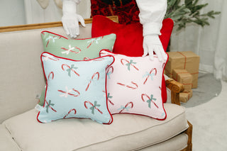 A person in white gloves arranges festive throw pillows, including the Childrens Green Candy Cane Pillow, on a beige sofa. Wrapped gifts and a decorated tree complete the cozy holiday decor in the background.