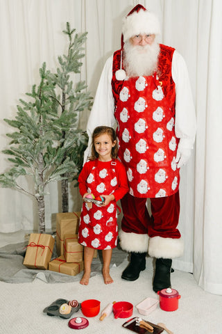A young girl and a man, both wearing matching Childrens Santa Aprons, stand by a decorated tree and gifts with toy baking supplies on the floor in front of them.