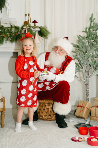 A smiling young girl wearing a Childrens Santa Apron stands next to Santa Claus, who is seated on a wicker stool with a bowl amid Christmas decorations and wrapped gifts.