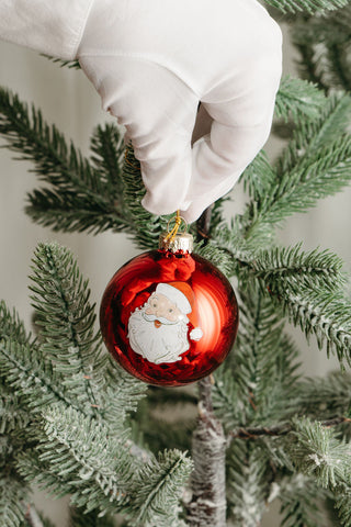 A hand in a white glove hangs a shiny red Santa Globe Ornament from the Santa Globe Ornaments set on a green Christmas tree, bringing festive cheer to the holiday scene.