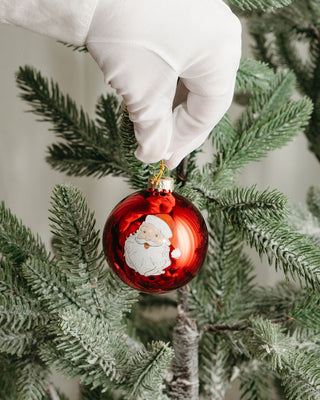 A gloved hand places a shiny red ornament from the Globe Ornament Variety 4 Pack, featuring a Santa Claus illustration, onto a green pine tree branch.