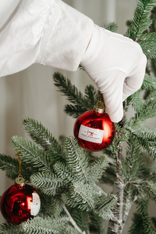 Wearing a white glove, someone hangs a red ornament on a green tree, while a Santa Globe Ornament (Single, 2 or 4 pack) sparkles on a lower branch, enhancing the coastal cheer of the festive scene.